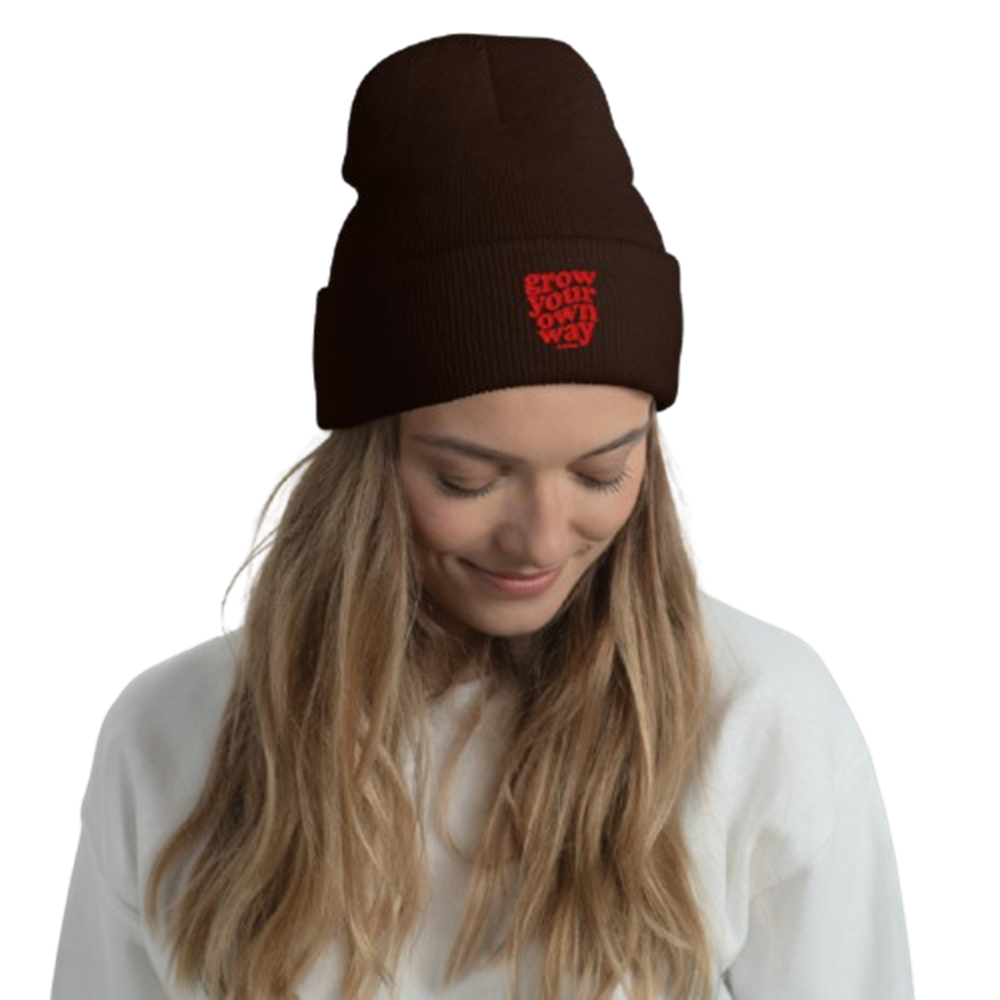 A woman wearing a dark brown knit beanie with a red embroidered logo that reads 'GROW YOUR OWN WAY'. The photo is taken against a clean white background.