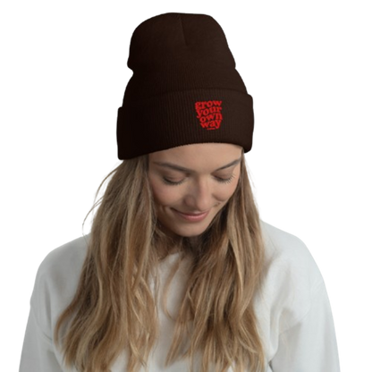 A woman wearing a dark brown knit beanie with a red embroidered logo that reads 'GROW YOUR OWN WAY'. The photo is taken against a clean white background.