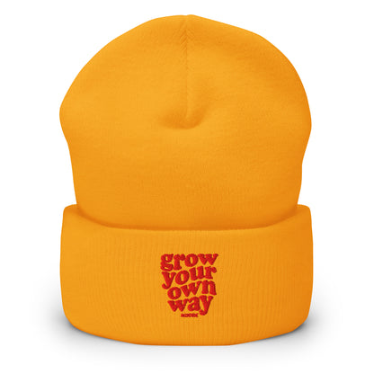 A bright orange knit beanie hat with a folded cuff, featuring the phrase 'grow your own way' embroidered in red on the front cuff.