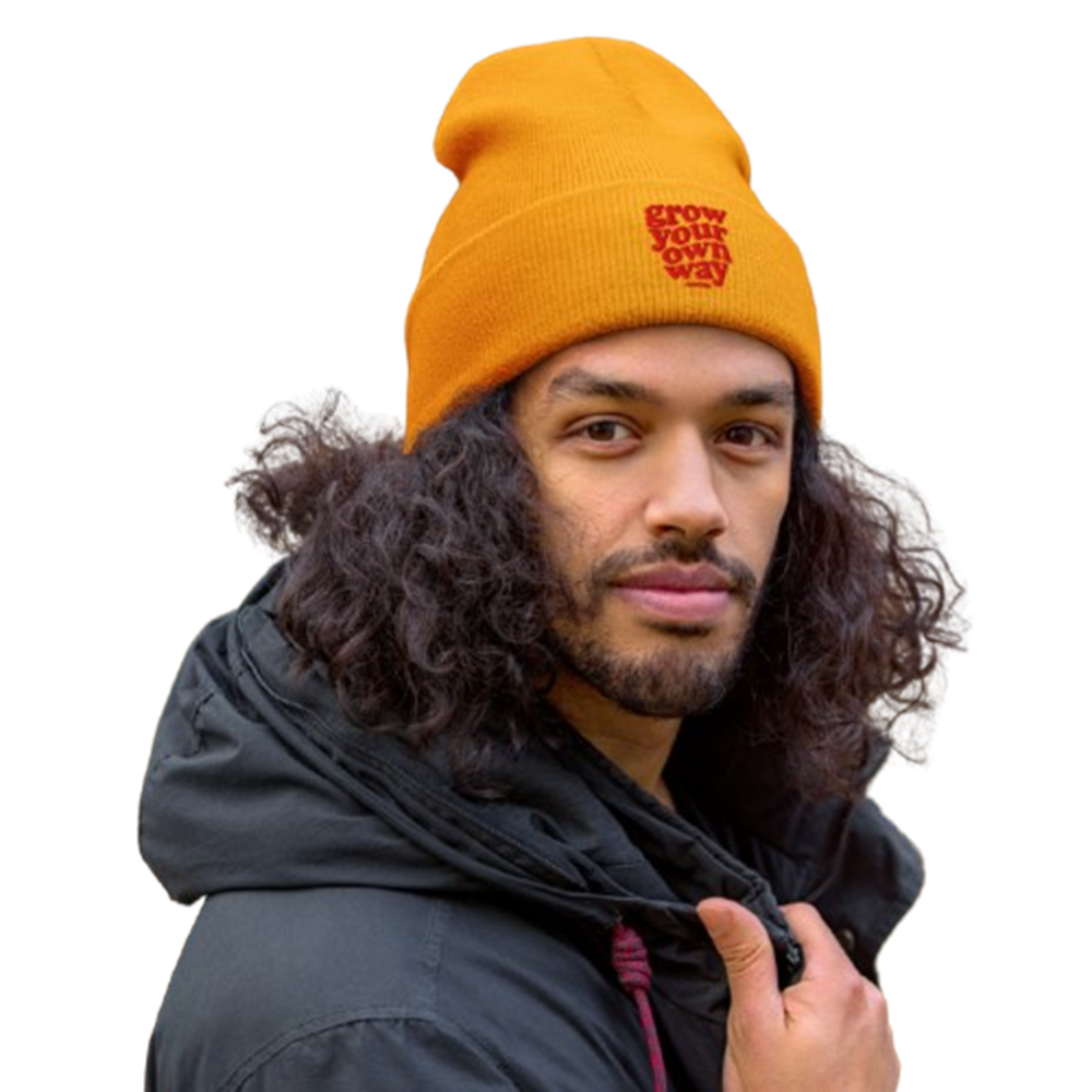 A man with long curly dark hair wearing a bright orange knit beanie with a red embroidered logo that reads 'GROW OUR OWN WAY'. He is also wearing a black hooded jacket against a plain white background.