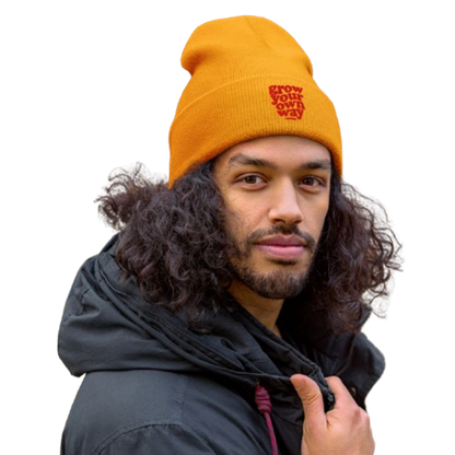 A man with long curly dark hair wearing a bright orange knit beanie with a red embroidered logo that reads 'GROW OUR OWN WAY'. He is also wearing a black hooded jacket against a plain white background.