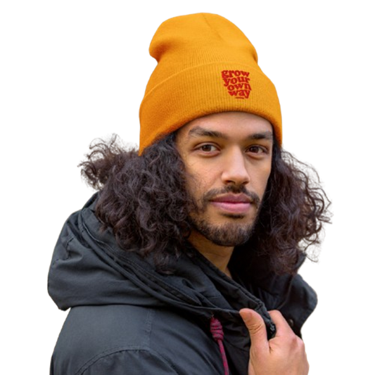 A man with long curly dark hair wearing a bright orange knit beanie with a red embroidered logo that reads 'GROW OUR OWN WAY'. He is also wearing a black hooded jacket against a plain white background.