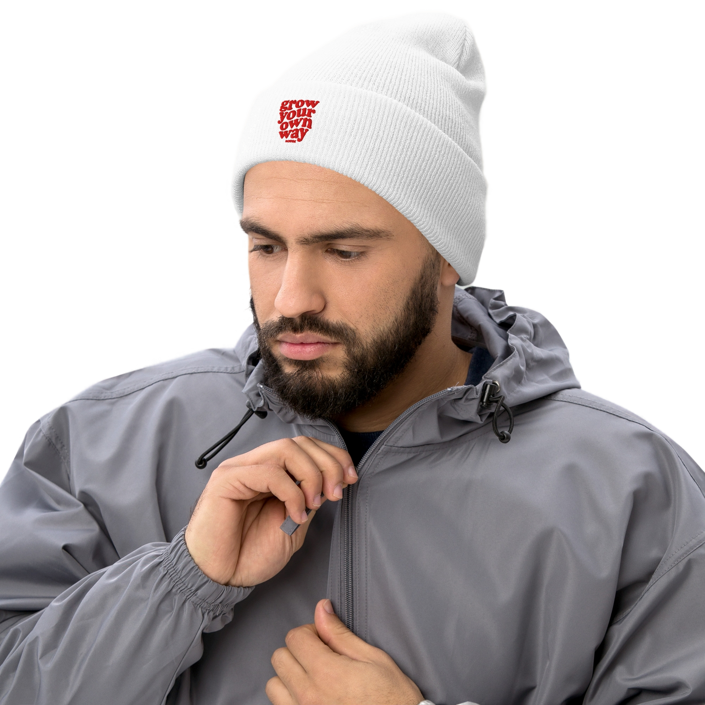 A smiling woman wearing a white knit beanie with a red embroidered logo, a black quilted vest over a gray shirt, leaning on a white table against a white background.