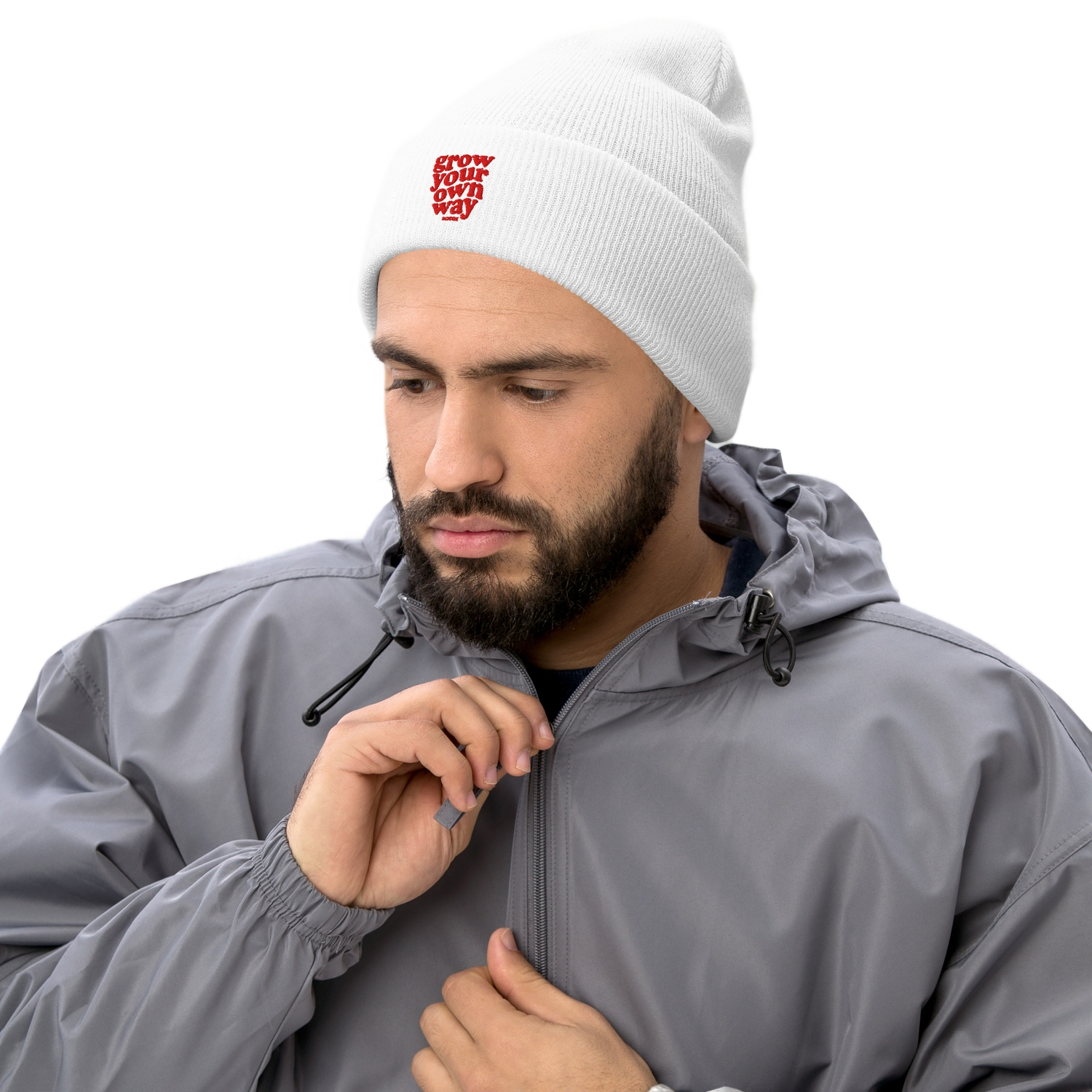 A smiling woman wearing a white knit beanie with a red embroidered logo, a black quilted vest over a gray shirt, leaning on a white table against a white background.