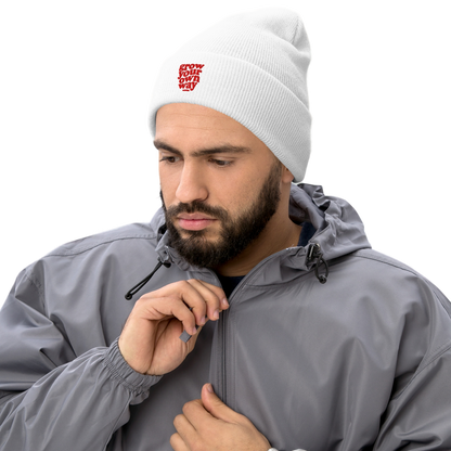 A smiling woman wearing a white knit beanie with a red embroidered logo, a black quilted vest over a gray shirt, leaning on a white table against a white background.