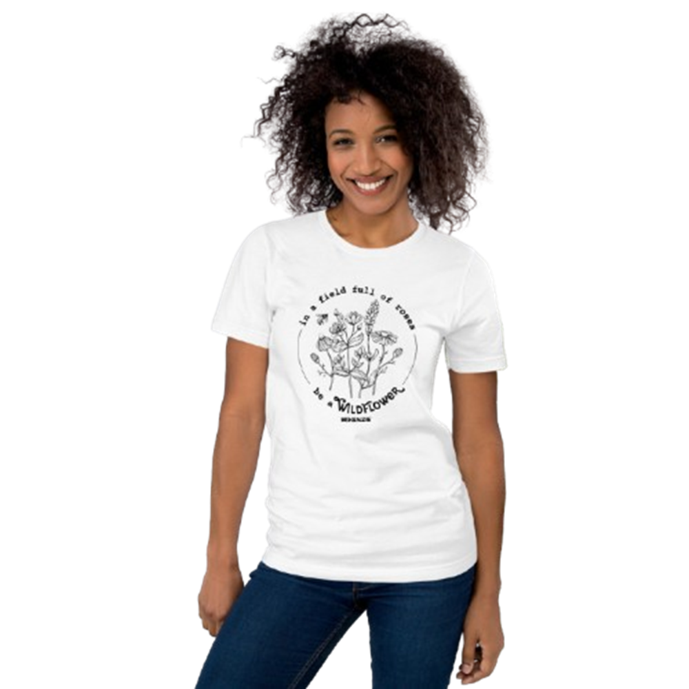 A smiling woman with curly dark hair wearing a white short-sleeve graphic T-shirt and blue jeans, standing against a plain white background.