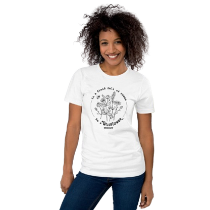 A smiling woman with curly dark hair wearing a white short-sleeve graphic T-shirt and blue jeans, standing against a plain white background.