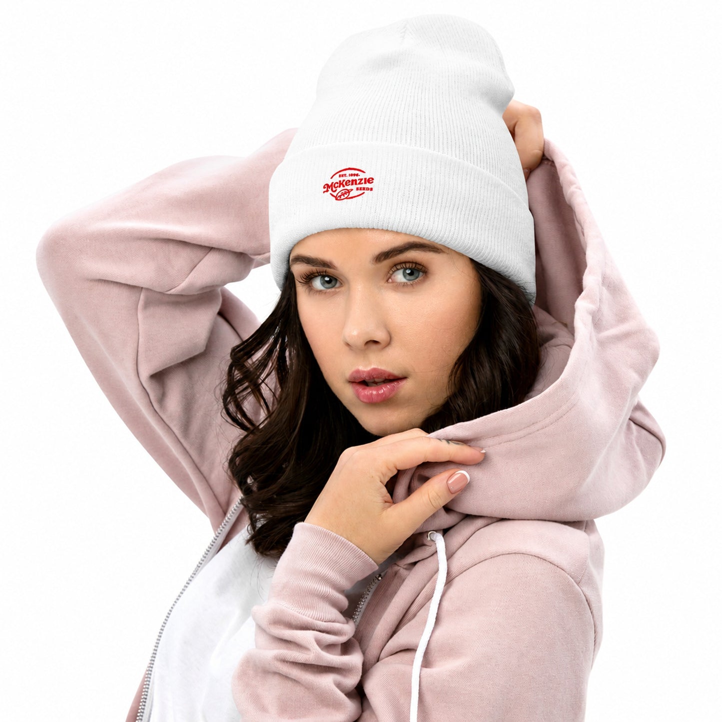 Woman wearing a light pink hoodie and white beanie with a logo on a gray background